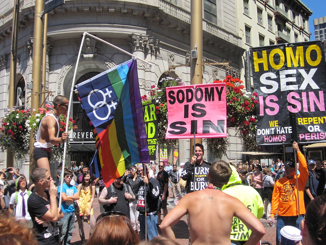 A line of counter-protesters stands opposite marchers during a Pride parade, with some holding signs and banners in opposition / Photo credit: Wikimedia Commons contributor