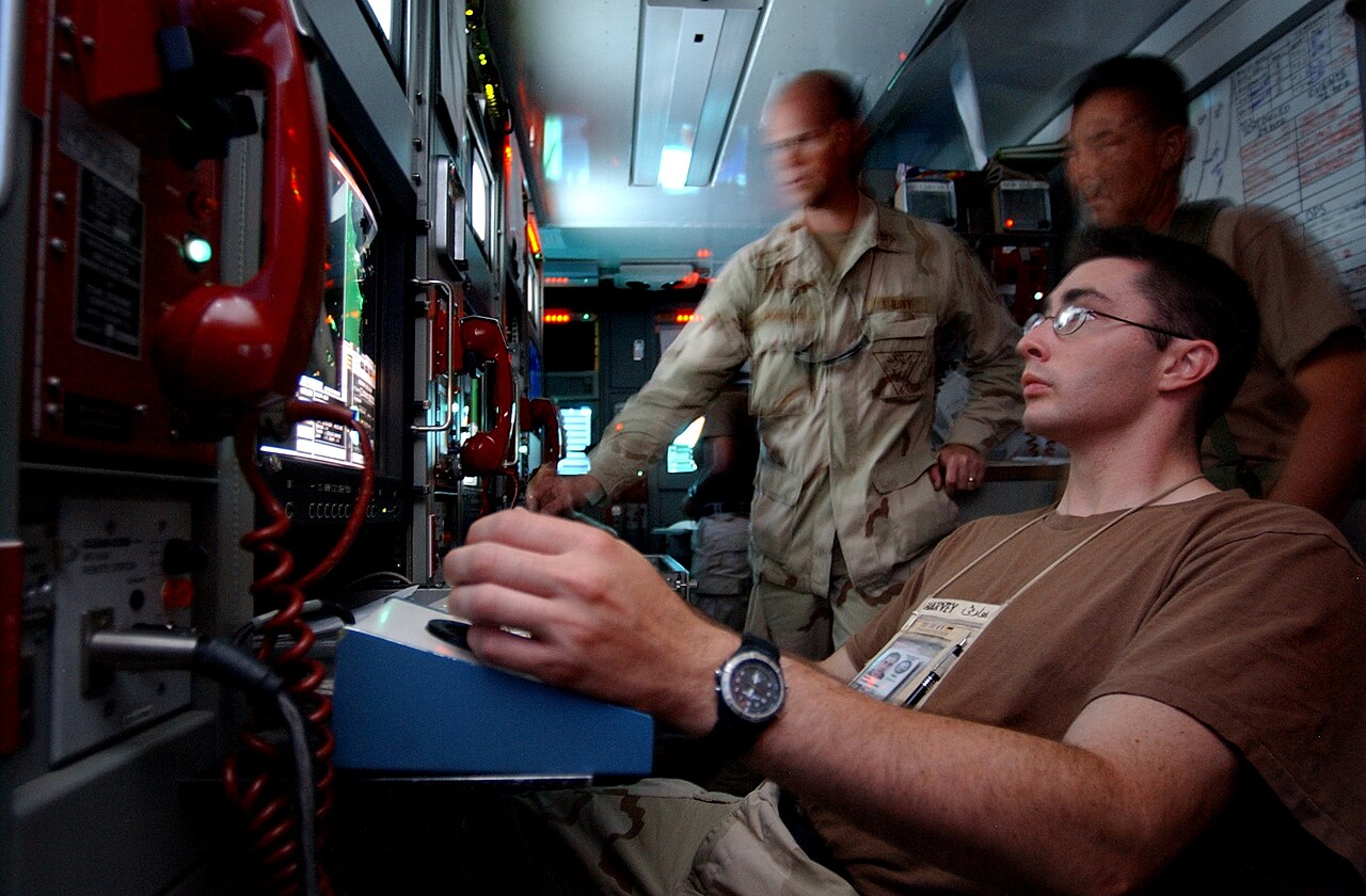 A military operator monitoring surveillance equipment in a dimly lit control room with multiple screens / Photo by PH1 Shane T. McCoy, U.S. Navy, released to the public domain
