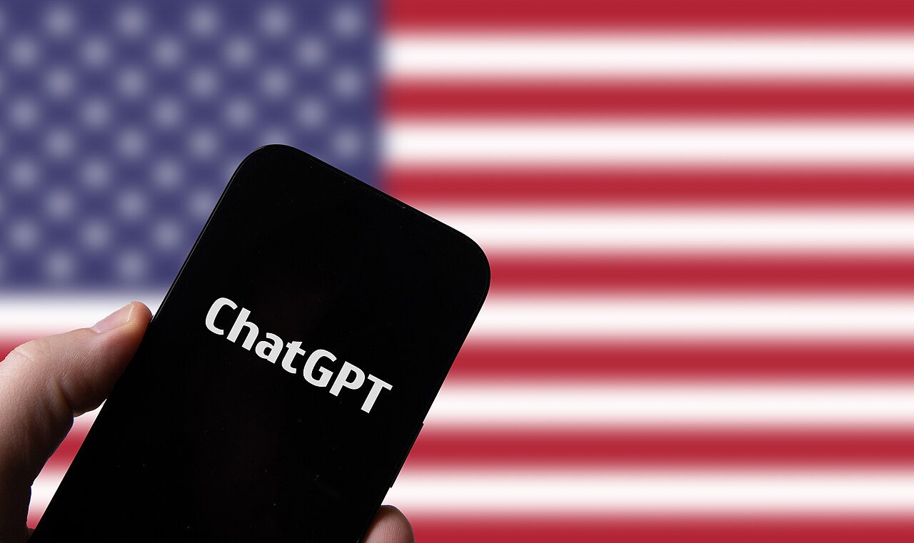 Hand holding a smartphone displaying the OpenAI ChatGPT interface against a U.S. flag background / Wikimedia Commons user